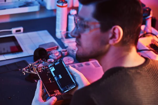 O reparador em óculos segurando um smartphone danificado, olhando para os lados em uma oficina de reparação moderna. Iluminação com luzes vermelhas e azuis — Fotografia de Stock