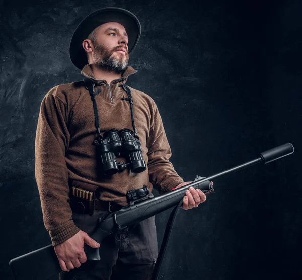 Mature hunter with rifle and binoculars. Studio photo against dark wall background — Stock Photo, Image