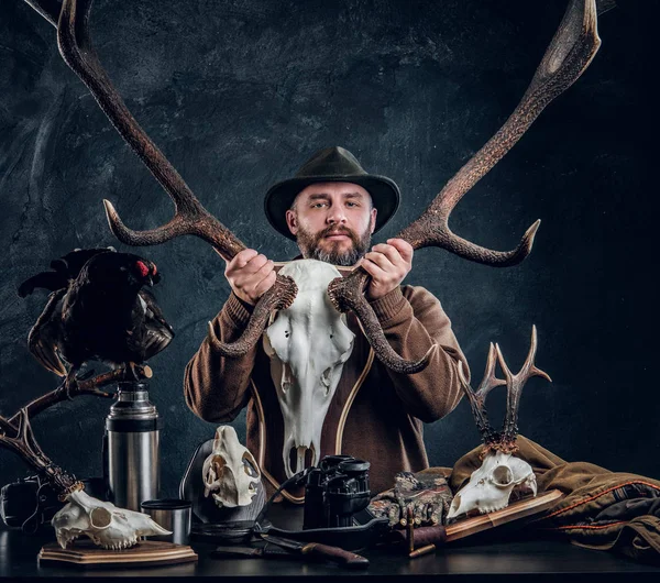 Reifer bärtiger Jäger mit Hut, der einen großen Hirschschädel hält, während er neben einem Tisch mit Ausrüstung und Trophäen steht. — Stockfoto