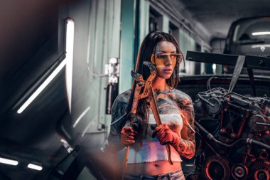 Dövmeli gövdeli hidrolik vinç atölye üzerinde askıya bir araba motoru yanındaki büyük bir İngiliz anahtarı ile poz koruyucu gözlük takan kadın modeli.