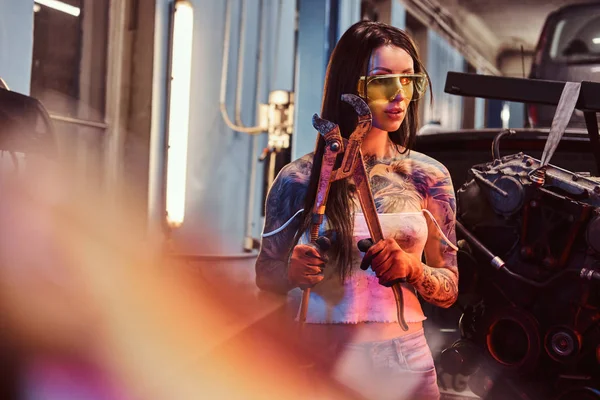 Female model with tattooed body wearing protective goggles posing with a big wrench next to a car engine suspended on a hydraulic hoist in the workshop.
