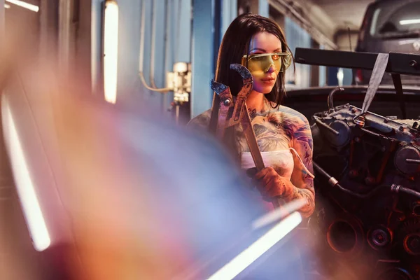 Modelki z tatuażem ciało noszenia okularów ochronnych, stwarzające duże kluczem płaskim obok silnika samochodu, zawieszone na podnośniku hydraulicznym w warsztacie. — Zdjęcie stockowe