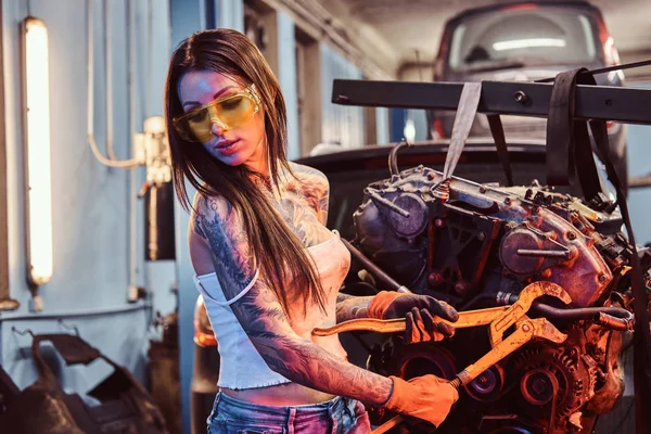 Stylish girl with tattooed body wearing protective goggles working with a car engine suspended on a hydraulic hoist in the workshop. — Stock Photo, Image