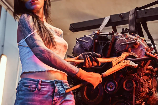 Elegante ragazza con corpo tatuato indossando occhiali protettivi che lavorano con un motore di auto sospeso su un paranco idraulico in officina . — Foto Stock