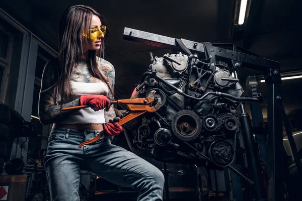 Stylish female model repairs a car engine suspended on a hydraulic hoist in the workshop. — Stock Photo, Image