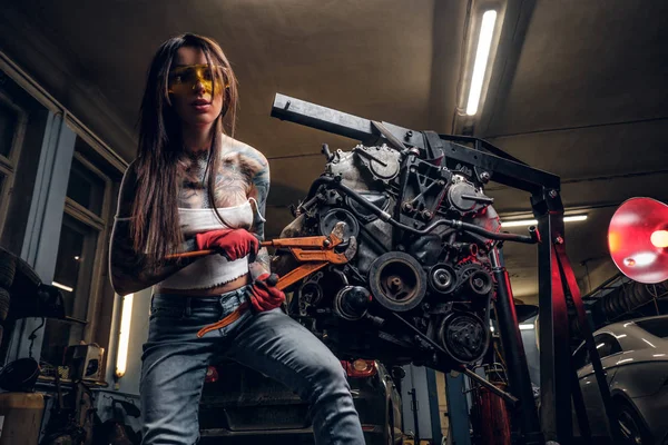 Modelo feminino elegante repara um motor de carro suspenso em um guincho hidráulico na oficina . — Fotografia de Stock