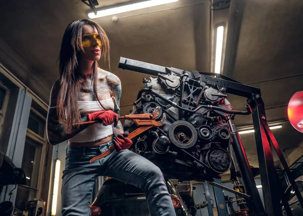 Elegante modello femminile ripara un motore di auto sospeso su un paranco idraulico in officina . — Foto Stock