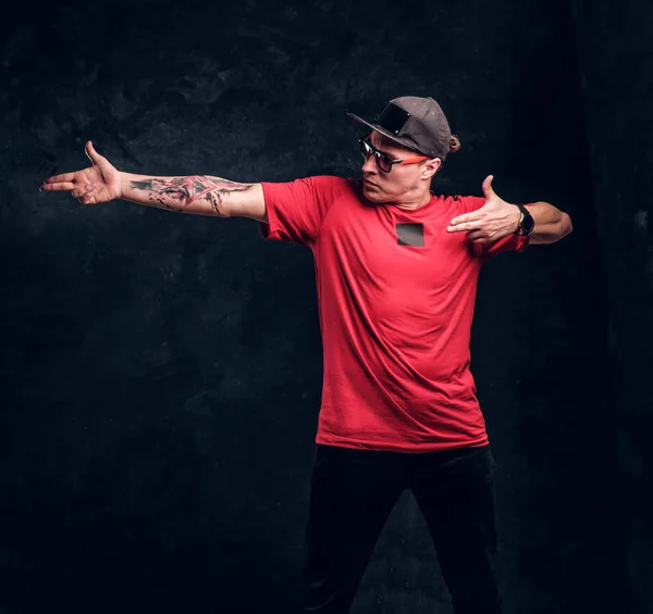 Elegáns férfi egy tetoválás a kezében öltözött hip-hop stílusban, pózol részére a fényképezőgép. Stúdió fotó egy sötét színű fallal szemben — Stock Fotó