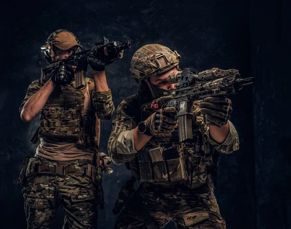 Dois soldados das forças especiais em equipamento de proteção total segurando rifles de assalto e apontando para os alvos. Foto do estúdio contra uma parede de textura escura . — Fotografia de Stock