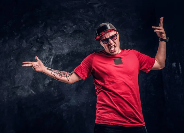 Elegante hombre con un tatuaje en la mano vestido con un estilo hip-hop, divirtiéndose y posando para una cámara. Foto del estudio contra una pared oscura — Foto de Stock
