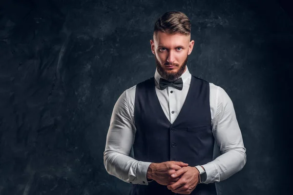 Elegant geklede bebaarde hipster in een vest met strikje. Studio foto tegen een donkere muur achtergrond — Stockfoto