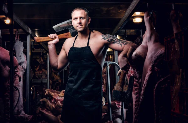 Sem camisa tatuado açougueiro em avental posando com machado em um armazém refrigerado no meio de carcaças de carne — Fotografia de Stock