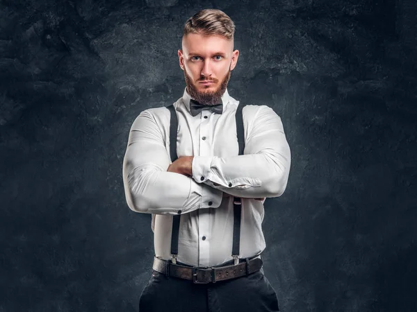 Stylishly dressed young man in shirt with bow tie and suspenders posing with his arms crossed and looking at a camera. Studio photo against dark wall background — Stock Photo, Image