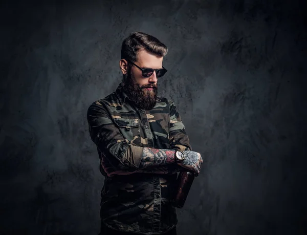 Un tipo hipster barbudo con camisa militar y gafas de sol sosteniendo una cerveza artesanal y mirando hacia abajo con una mirada pensativa. Estudio foto contra la pared oscura —  Fotos de Stock