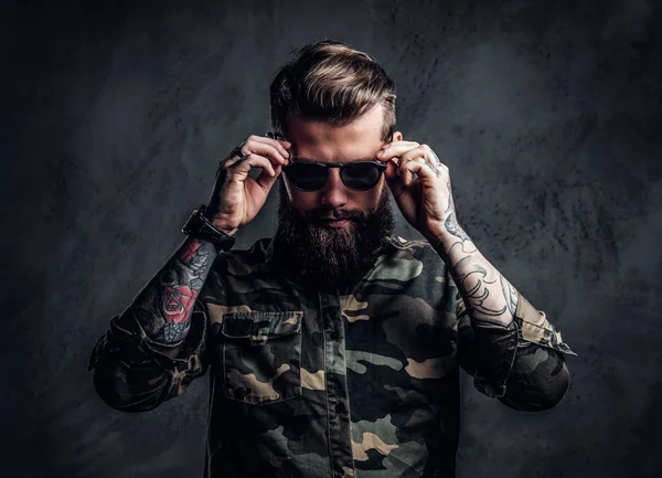 Portrait of a stylish bearded guy with tattooed hands in the military shirt holding hands on sunglasses. Studio photo against dark wall — Stock Photo, Image