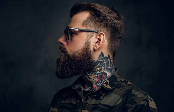 Profile of a stylish bearded guy with tattooed hands in the military shirt. Studio photo against dark wall — Stock Photo, Image