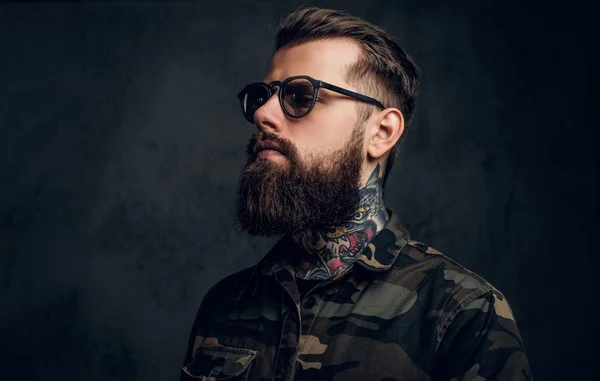 Closeup portrait of a bearded man with a tattoo on his neck in sunglasses wearing a military shirt. Studio photo against dark wall — Stock Photo, Image