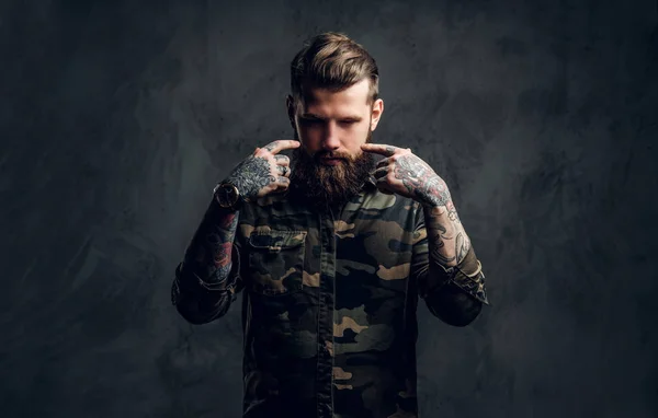 Ritratto di un ragazzo barbuto elegante con le mani tatuate nella camicia militare. Studio foto contro muro scuro — Foto Stock