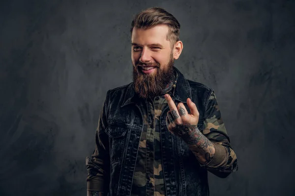 A crazy bearded man in the military shirt and denim vest with tattooed hands showing the fuck sign. Studio photo against dark wall