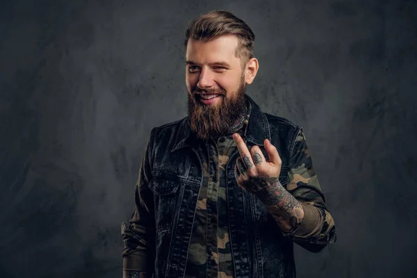 En galen skäggiga man i den militära skjortan och denim väst med tatuerade händer visar fan tecken. Studio Foto mot mörk vägg — Stockfoto