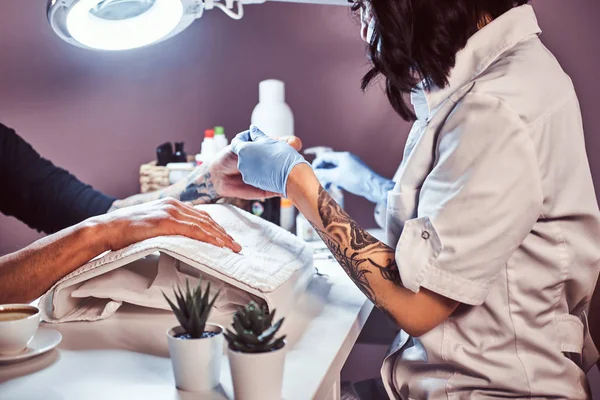 Um homem com uma tatuagem na mão a receber uma manicura de um esteticista no salão de beleza. Mãos fechadas — Fotografia de Stock