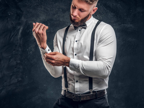 Stylishly dressed young man in shirt with bow tie and suspenders. Studio photo against dark wall background