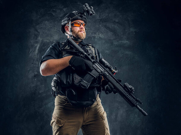 Special forces soldier wearing body armor and helmet with night vision holding an assault rifle. Studio photo against a dark textured wall