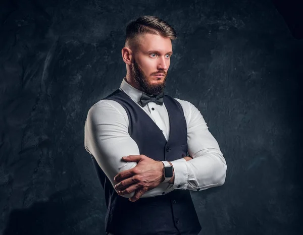 Retrato de um jovem elegantemente vestido em um colete com laço posando com os braços cruzados e olhando para o lado. Foto do estúdio contra fundo de parede escura — Fotografia de Stock