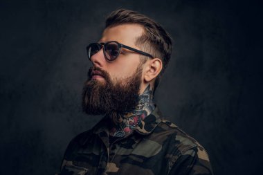 Askeri bir gömlek giyen güneş gözlüğü boynunda bir dövme ile sakallı bir adamın closeup portre. Koyu duvara karşı stüdyo fotoğrafı