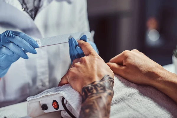 Een man met een tatoeage op zijn hand ontvangt een manicure door een schoonheidsspecialiste in de schoonheidssalon. Close-up handen — Stockfoto