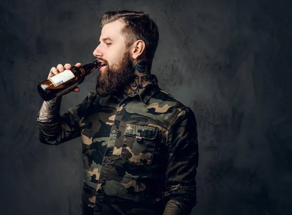 Elegante chico hipster barbudo en camisa militar disfruta del sabor de la cerveza artesanal. Estudio foto contra la pared oscura —  Fotos de Stock