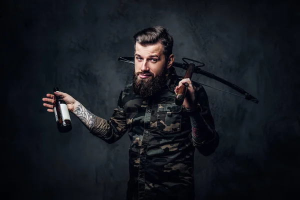 Elegante hombre hipster barbudo con camisa militar sosteniendo una cerveza artesanal y una ballesta medieval. Estudio foto contra la pared oscura — Foto de Stock
