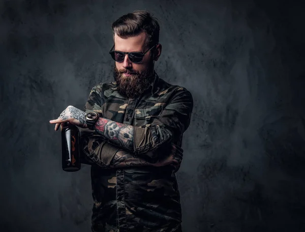 Un tipo hipster barbudo con camisa militar y gafas de sol sosteniendo una cerveza artesanal y mirando hacia abajo con una mirada pensativa. Estudio foto contra la pared oscura —  Fotos de Stock