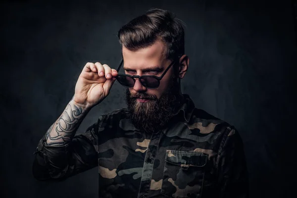 Retrato de um cara barbudo elegante com as mãos tatuadas na camisa militar segurando a mão em óculos de sol e olhando para uma câmera. Estúdio foto contra parede escura — Fotografia de Stock