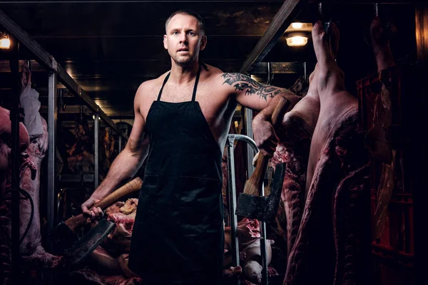 Shirtless tattooed butcher in apron posing with ax in a refrigerated warehouse in the midst of meat carcasses — Stock Photo, Image