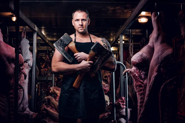 Carnicero tatuado sin camisa en delantal posando con hacha en un almacén refrigerado en medio de cadáveres de carne — Foto de Stock