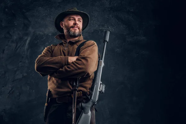 Portrait of a bearded hunter with rifle posing with his arms crossed. Studio photo against a dark wall background — Stock Photo, Image
