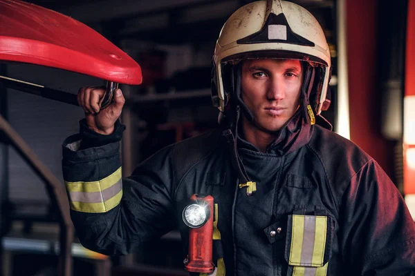 Retrato de un guapo bombero vestido con un uniforme protector con linterna incluido de pie en un garaje de bomberos —  Fotos de Stock