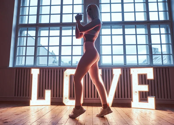 Schöne Fitness-Model in Sportbekleidung posiert in der Nähe von großen beleuchteten Wort Liebe in einem Raum mit Lift-Interieur. Liebe, Valentinstag, Schönheitskonzept — Stockfoto