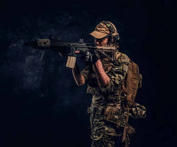 L'unité d'élite, soldat des forces spéciales en uniforme de camouflage tenant un fusil d'assaut avec une vue laser et vise la cible. Studio photo contre un mur sombre — Photo