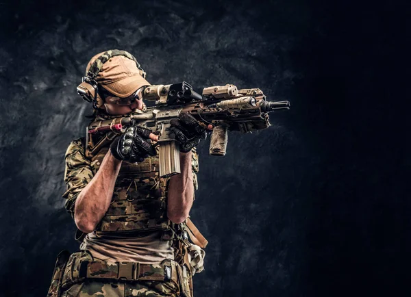 A unidade de elite, soldado das forças especiais em uniforme de camuflagem segurando um rifle de assalto com uma mira de laser e aponta para o alvo. Foto de estúdio contra uma parede escura — Fotografia de Stock