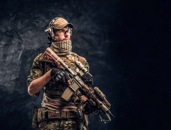 Повністю обладнаний солдат у камуфляжній формі, що тримає штурмову гвинтівку. Студійне фото на тлі темної стіни — стокове фото