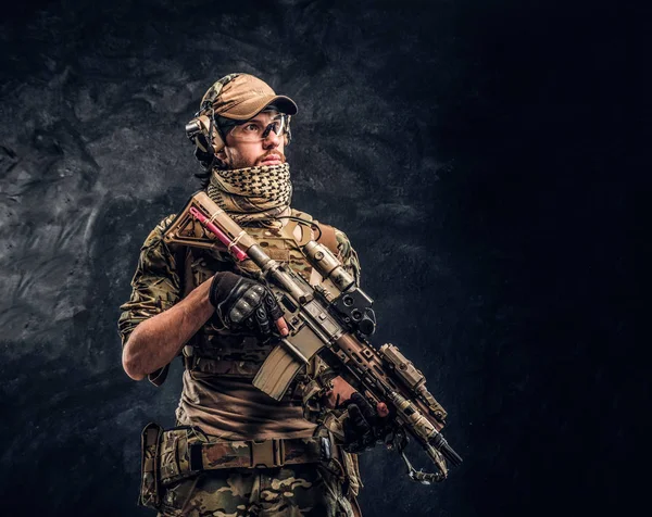 Soldat entièrement équipé en uniforme de camouflage tenant un fusil d'assaut. Studio photo contre un mur sombre — Photo