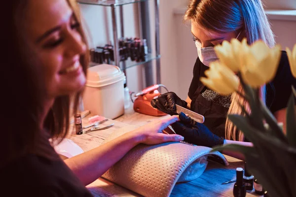 Nail master está trabalhando em unhas de cliente com arquivo de unhas. Mestre está usando máscara de proteção e luvas de látex preto . — Fotografia de Stock