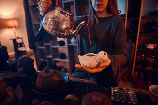 Oriental master wearing a gray dress holding a kettle and pouring natural tea into a teapot in the dark room with a wooden interior. Tradition, health, harmony. — Stock Photo, Image