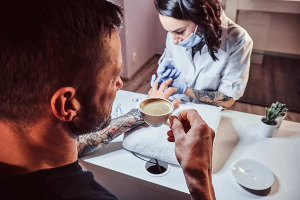 A man receiving a manicure in the beauty salon, relaxing and drinking a coffee while beautician master does manicure procedure — Stock Photo, Image