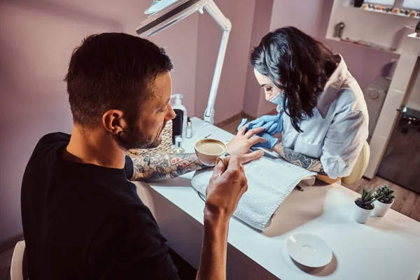 Um homem recebendo uma manicure no salão de beleza, relaxando e bebendo um café enquanto o mestre esteticista faz o procedimento de manicure — Fotografia de Stock