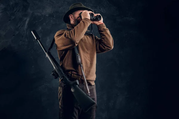 Hunter with shotgun watching through binoculars. Studio photo against dark wall background — Stock Photo, Image