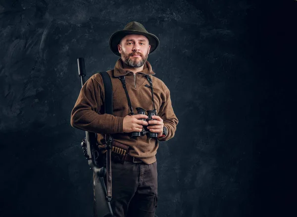 Mature hunter with rifle holding binoculars and looking at a camera. Studio photo against dark wall background — Stock Photo, Image
