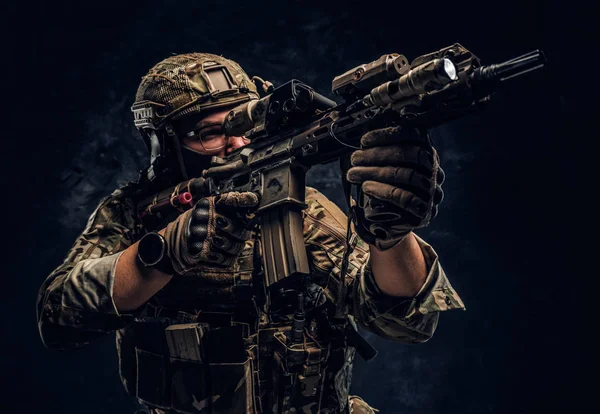 Close-up studio photo against a dark wall. The elite unit, special forces soldier in camouflage uniform holding an assault rifle with a laser sight and aims at the targe — Stock Photo, Image